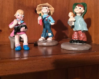 May include: Three ceramic figurines of children on a wooden shelf. The figurine on the left is a girl wearing a pink dress and holding a black and white dog. The figurine in the middle is a boy wearing overalls and a straw hat, holding a carrot. The figurine on the right is a boy wearing a blue shirt and a green hat, holding a doll.