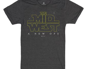 Puede incluir: Una camiseta gris oscuro con un gráfico amarillo de "The Mid West A New Hope" sobre un fondo de campo de estrellas.