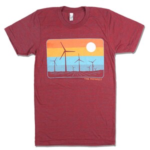 Op de afbeelding: Rood T-shirt met een afbeelding van windturbines in een veld met zonsondergang en de tekst "THE MIDWEST".