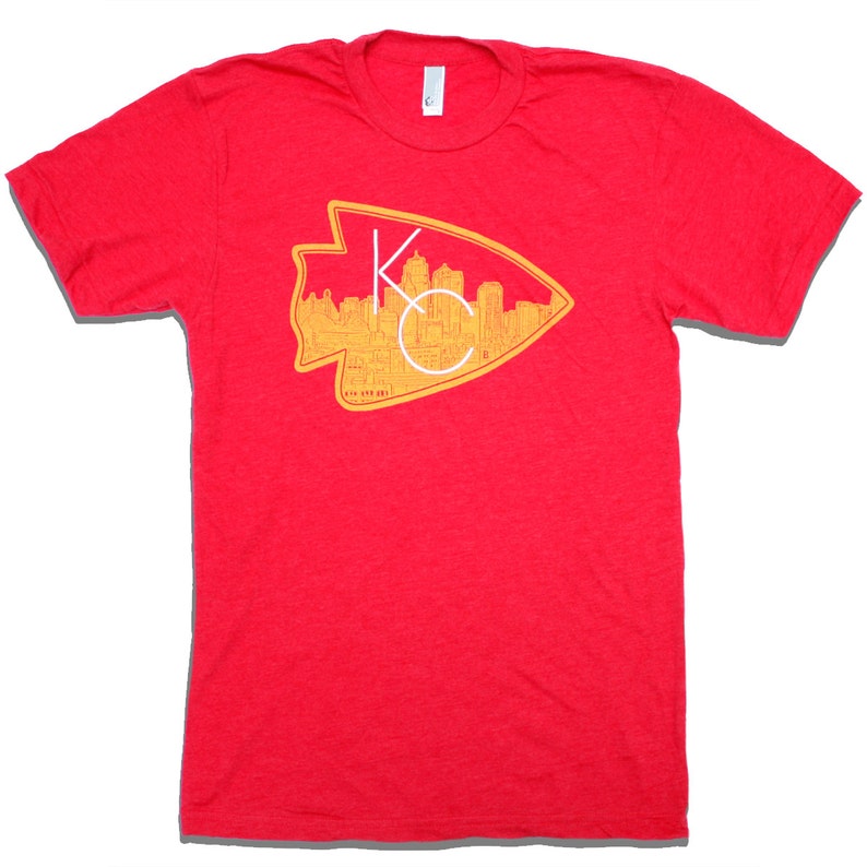 May include: Red t-shirt with a yellow and white graphic of the Kansas City skyline inside an arrowhead. The text "KC" is in white.