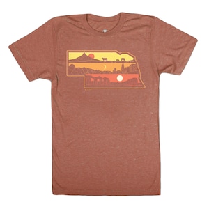 Op de afbeelding: Een maronkleurig T-shirt met een afbeelding van de staat Nebraska. De afbeelding is een silhouet van de staat met verschillende scènes die op elkaar zijn gelegd, waaronder een berg, een zonsondergang, een veld met koeien en een skyline van de stad.