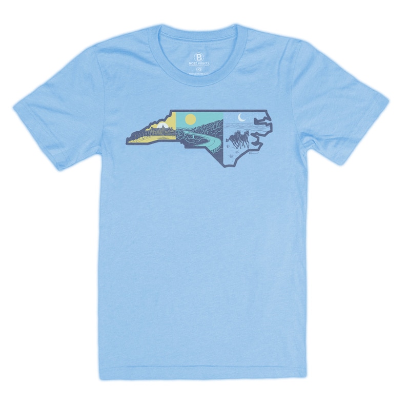 May include: Light blue t-shirt featuring a graphic of the state of North Carolina with different scenes from the state, including a mountain range, a sunset, a river, and horses running.