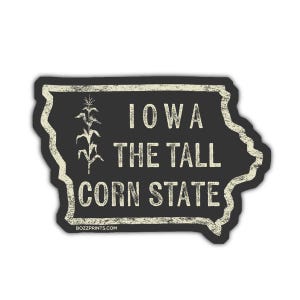 Könnte beinhalten: Ein schwarzer Aufkleber in Form von Iowa mit einem weißen, abgenutzten Rand. Der Text "IOWA THE TALL CORN STATE" ist in Weiß gedruckt, zusammen mit einer Maiskolben-Grafik. Die Website "bozzprints.com" ist unten aufgedruckt.