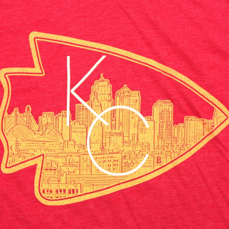 May include: A red t-shirt with a gold outline of the Kansas City skyline inside an arrowhead. The skyline includes the words "KC" in white.