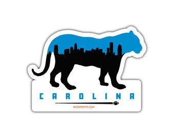 May include: A black silhouette of a panther with a blue cityscape inside. The text "CAROLINA" is below the panther with a football and a line underneath. The text "BOZZPRINTS.COM" is below the line.