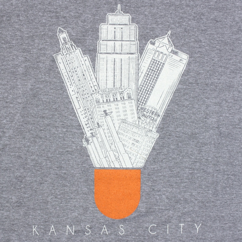 May include: A gray t-shirt with a white graphic of the Kansas City skyline in the shape of a shuttlecock. The shuttlecock is filled with an orange color. The text "KANSAS CITY" is printed below the graphic.