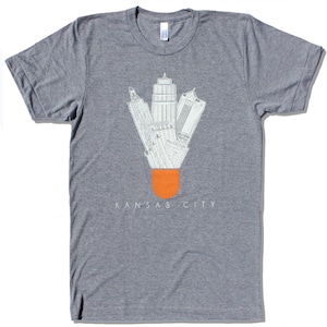 May include: A gray t-shirt with a white graphic of the Kansas City skyline designed to look like a shuttlecock. The text "KANSAS CITY" is printed below the graphic.