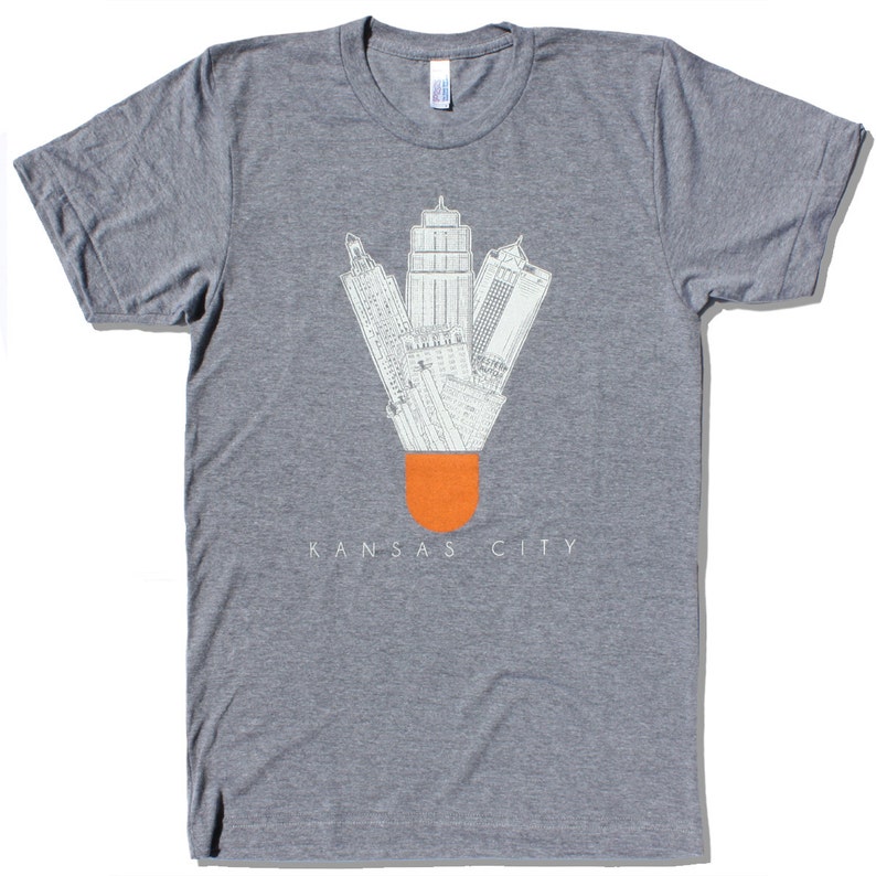 May include: A gray t-shirt with a white graphic of the Kansas City skyline designed to look like a shuttlecock. The text "KANSAS CITY" is printed below the graphic.