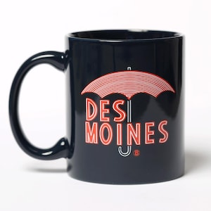 以下が含まれることがあります： 大きなハンドルが付いた濃い青色のセラミックマグカップ。「DES MOINES」の文字が赤と白で表示され、その上に赤と白の傘のグラフィックが描かれています。