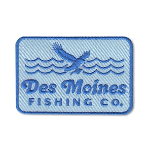 Könnte beinhalten: Rechteckiger Aufnäher mit hellblauem Hintergrund, der einen blauen Adler über blauen Wellen zeigt. Der Text "Des Moines FISHING CO." ist in Blau mit einer stilisierten Schriftart gestickt. Der Aufnäher hat einen hellblauen Rand.