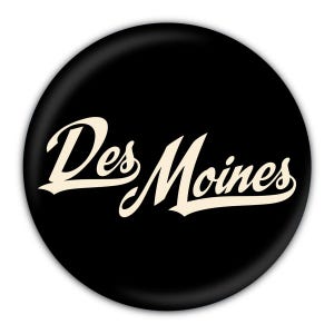 May include: A black round button with the words "Des Moines" in a cream-colored, cursive font. The text is centered on the button, and the overall design is simple and clean.