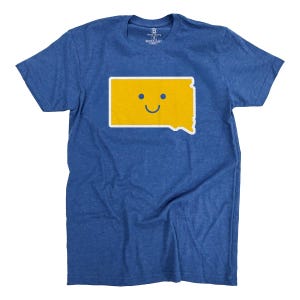 Peut inclure: T-shirt bleu avec un contour jaune de l'État du Dakota du Sud et un visage souriant. Le motif est centré sur le t-shirt. Le t-shirt est fait d'une matière douce.