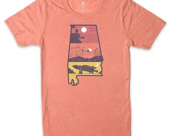 May include: A coral-colored t-shirt with a graphic of the state of Alabama. The graphic features three scenes: a couple standing on a cliff overlooking a sunset, a tent on a lake with tall grass, and a rocket launching with a crescent moon in the sky.
