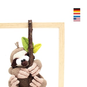 Puede incluir: Un juguete de peluche de perezoso tejido a crochet que sostiene una rama con hojas verdes. El perezoso es marrón y tiene una cara blanca con ojos negros. El juguete está sentado en un marco de madera.