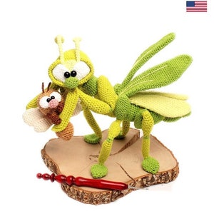 Puede incluir: Una mantis religiosa de crochet verde sosteniendo un insecto de crochet marrón más pequeño. La mantis tiene ojos grandes y redondos y está de pie sobre una base de madera. La base tiene un gancho de crochet rojo en ella.