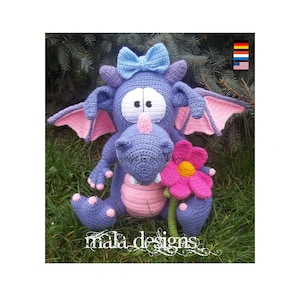 Può includere: Un drago di uncinetto viola con ali rosa e un fiore rosa. Il drago ha un fiocco blu sulla testa e grandi occhi bianchi. Il drago è seduto su un letto di erba verde.