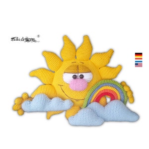 Op de afbeelding: Een gehaakte zon met een glimlach, een regenboog en twee wolken. De zon is geel, de regenboog is veelkleurig en de wolken zijn lichtblauw.