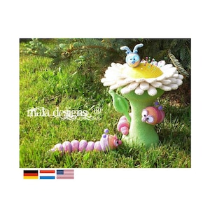 Könnte beinhalten: Ein gehäkeltes Gänseblümchen-Nähkissen mit einer blauen und weißen gehäkelten Wanze oben drauf. Das Nähkissen sitzt auf einem grünen gehäkelten Stiel mit zwei rosa und lila gehäkelten Raupen.