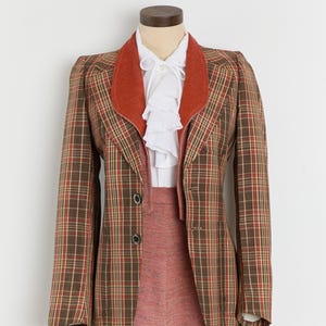 May include: A vintage plaid blazer with a rust-colored collar and a matching skirt. The blazer features a brown, red, and tan plaid pattern. A white blouse with ruffled detailing is visible beneath the blazer. The skirt is a solid, muted red color.