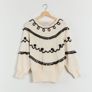 Puede incluir: Suéter color crema con ribete decorativo negro. El suéter presenta cuello redondo y mangas largas. El ribete negro incluye diseños de bucles, líneas onduladas y pompones. El suéter está colgado en una percha de madera.