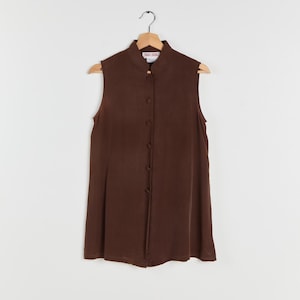 May include: A sleeveless, button-down top in a rich brown hue. The top features a mandarin collar and a series of buttons down the front. It hangs on a wooden hanger against a plain white background. The top is a solid color and appears to be made of a smooth fabric.