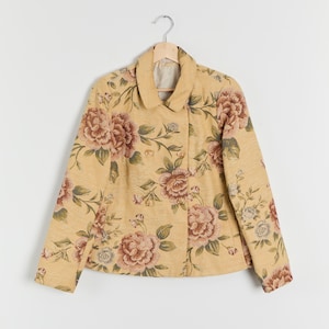 May include: A double-breasted jacket with a floral pattern. The jacket is a light yellow color with large, detailed flowers in shades of pink, brown, and green. The jacket has a collared neckline and is hanging on a wooden hanger.