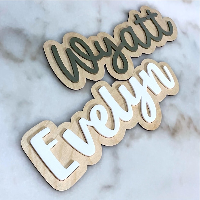May include: Two custom name signs crafted from light-coloured wood with a layered design. The top sign features the name Wyatt in dark olive green script, while the bottom sign displays the name Evelyn in white script, both mounted on wood backings.