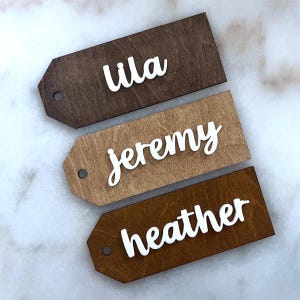 May include: Three wooden name tags with a tag shape. Each tag has a different wood stain color and a white cursive name: Lila, Jeremy, and Heather. Each tag has a small hole at the top for hanging.