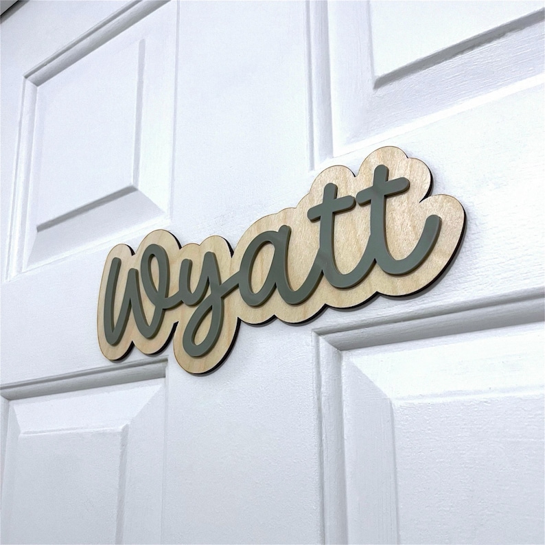 May include: A custom name sign featuring the name Wyatt in dark grey script lettering. The letters are mounted onto a light-coloured wooden backing with a scalloped edge, creating a layered effect that stands out against the white door surface.