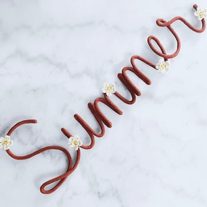 May include: The word "Summer" is spelled out in brown rope with white crocheted flowers attached to each letter.
