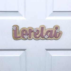 May include: A custom wooden name sign featuring the name Lorelai in a cursive script. The letters are a muted mauve colour, layered over a light wood-toned backing. Small white flowers with yellow centres are attached to the sign as accents.