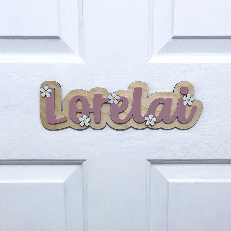 May include: A custom wooden name sign featuring the name Lorelai in a cursive script. The letters are a muted mauve colour, layered over a light wood-toned backing. Small white flowers with yellow centres are attached to the sign as accents.