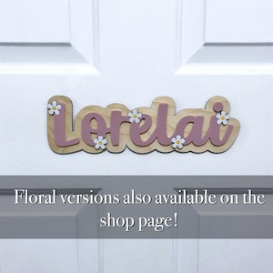 May include: A wooden name sign featuring the name Lorelai in mauve script lettering mounted on a light wood backing. The sign is decorated with small white flowers with yellow centres. The text Floral versions also available on the shop page! is visible.