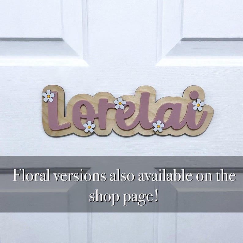 May include: A wooden name sign featuring the name Lorelai in mauve script lettering mounted on a light wood backing. The sign is decorated with small white flowers with yellow centres. The text Floral versions also available on the shop page! is visible.