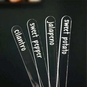 May include: Four clear acrylic garden markers with rounded tops. Each marker has white text: cilantro, sweet pepper, jalapeno, and sweet potato. The markers are arranged at an angle against a dark background.