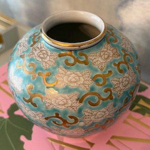 Puede incluir: Un jarrón de cerámica redondo con fondo turquesa y motivos florales blancos. Acentos y adornos dorados adornan el jarrón, que mide aproximadamente 15 cm de diámetro. El jarrón tiene un interior blanco y un borde dorado.