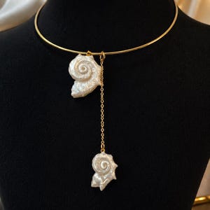 May include: A gold-toned metal choker necklace featuring two white, iridescent, spiral-shaped shell pendants. One shell is attached directly to the thin, circular gold collar, while the other hangs from a delicate gold chain drop.