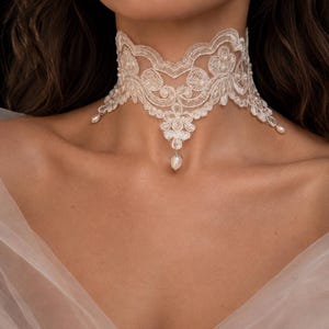 May include: An ivory lace choker necklace featuring intricate floral embroidery and scalloped edges. The design is embellished with small, round white pearls integrated into the lace pattern and finished with three dangling pearl drops at the center and sides.