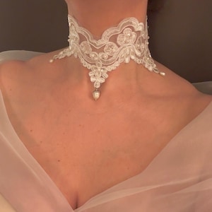 May include: A delicate white lace choker necklace with floral and pearl accents. The intricate design features scalloped edges and a central pendant with a teardrop-shaped pearl. The necklace is designed to be worn around the neck.