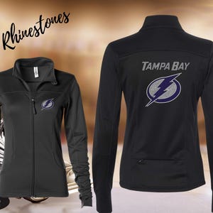 May include: Black zip-up jacket with the Tampa Bay logo on the front and back. The logo is a lightning bolt with a purple and white design. The text "In Rhinestones" is above the jacket.