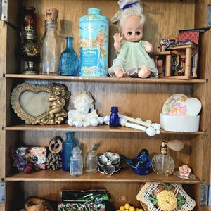 May include: A wooden cabinet with four shelves containing various vintage items including a small doll in a green dress, a blue floral tin, glass bottles, a gold heart-shaped frame with a cherub, a white ceramic angel, a blue glass bird, and yellow beads.