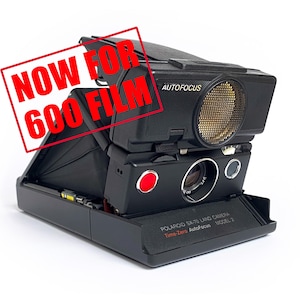 Peut inclure: Un appareil photo Polaroid SX-70 Land Camera modèle 1 Time-Zero Autofocus noir avec les mots "NOW FOR 600 FILM" en lettres rouges sur un fond blanc.