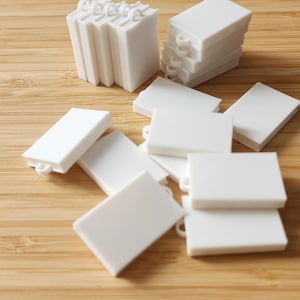 May include: A collection of small, white, rectangular objects with rounded corners and a loop on one side. Some are stacked, while others are scattered on a wooden surface. The objects resemble miniature books or tags.
