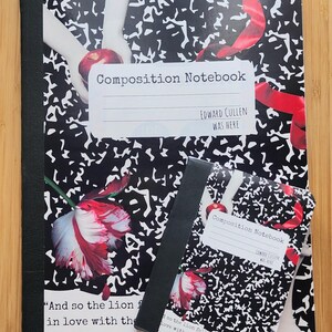 May include: Composition notebooks with black and white speckled covers. The larger notebook has a label that reads "Composition Notebook" and "EDWARD CULLEN WAS HERE." A smaller notebook is on top. The notebooks have images of a red apple and red flowers.