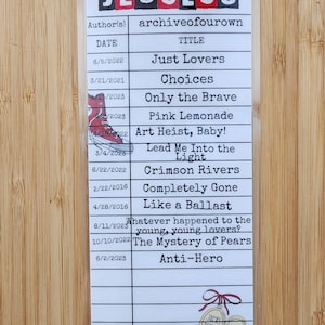 May include: A white bookmark with a list of titles and dates, including "Just Lovers" and "Anti-Hero." The word "JEGULUS" is at the top. A small illustration of a red shoe is present. The bookmark is on a wooden surface.