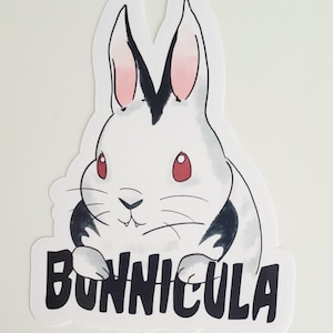 May include: A white rabbit with black ears and red eyes is shown with fangs. The rabbit is sitting with its paws in front of it. The text "BONNICULA" is written in black below the rabbit.