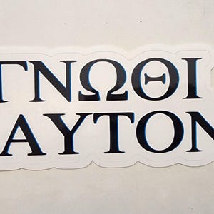 May include: A white sticker with a black border and Greek text. The text reads "ΓΝΩΘΙ ΣΑΥΤΟΝ" in a bold, sans-serif font. The sticker is rectangular with rounded corners.