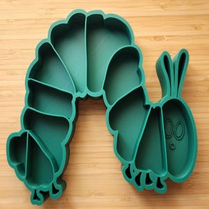 May include: A dark teal plastic tray shaped like a segmented caterpillar with a curved body. The tray features multiple internal compartments defined by raised walls, with a stylized face including two antennae and oval eyes on the right end.