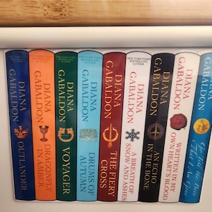 May include: A set of ten paperback books with blue, green, orange, and white covers. The books are all by Diana Gabaldon and feature the titles "Outlander", "Dragonfly in Amber", "Voyager", "Drums of Autumn", "The Fiery Cross", "A Breath of Snow and Ashes", "An Echo in the Bone", "Written in My Own Heart's Blood", "Go Tell the Bees That I Am Gone", and "The Fiery Cross".