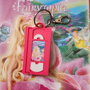 May include: A pink VHS tape-shaped keychain featuring a miniature Barbie Fairytopia movie cover. The keychain is attached to a silver keyring and clasp. The background shows the Barbie Fairytopia movie cover art with the text "Discover A Magical New Land".